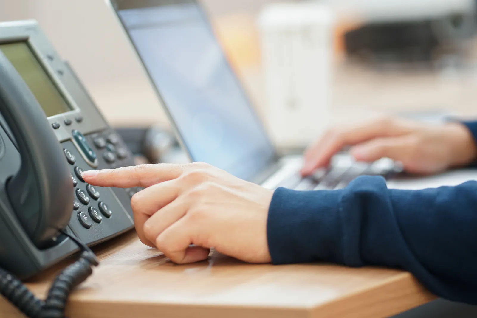 A hand dialing a desk phone beside a laptop.